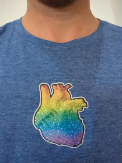 Close up of an anatomical heart overlayed with a rainbow gradient glitter printed on a blue t-shirt.