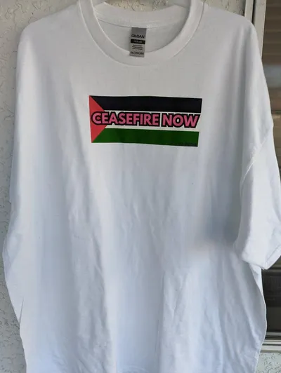 The flag of Palestine with the phrase Ceasefire Now written across it in pink letters, screen printed on a white t-shirt.