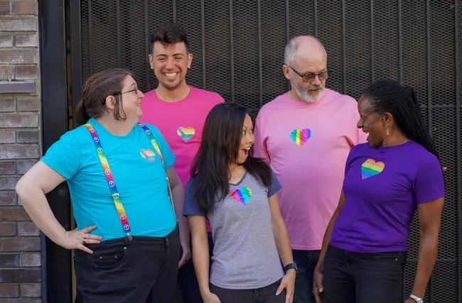 A group of five people wearing t-shirts looking at each other excitedly. Each person's t-shirt has a glitter design on it: the two on the left have the Progress Pride flag in a heart on the upper left chest, the two in the middle have vertical rainbow stripes in a heart in the center of the chest, and the one on the right has a classic pride flag in a heart in the center of the chest.