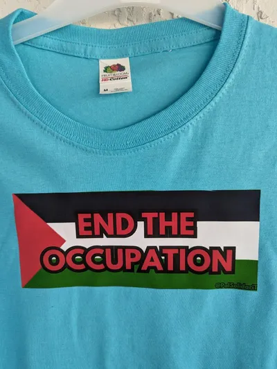 The flag of Palestine with the phrase End the Occupation written across it in red letters, screen printed on a light blue t-shirt.