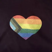 Close up of a Progress Pride flag in the shape of a heart printed on a black t-shirt.