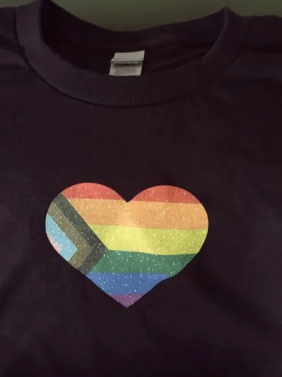 Close up of a Progress Pride flag in the shape of a heart printed on a black t-shirt.