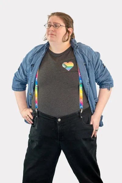 A Progress Pride flag in the shape of a heart glitter printed on the upper left side of a grey t-shirt, worn by a white person with rainbow suspenders.