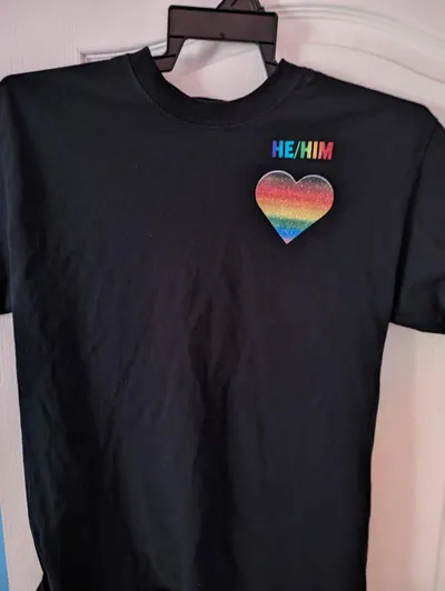 Rainbow gradient he/him pronouns with a left-to-right purple to red rainbow gradient above a heart with a top-to-bottom black-to-brown-to-rainbow gradient glitter printed on the upper left side of a black t-shirt.