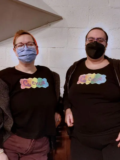 Two people wearing black shirts glitter printed with the words Q Choir in purple on a music staff over a rainbow background.