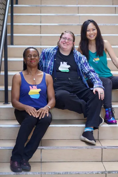 Three people seated on concrete stairs. The person at the top wears a teal tank top glitter printed with a Progress Pride flag in the shape of a heart. The person in the middle wears a black t-shirt glitter printed with a genderqueer flag in the shape of an anatomical heart. The person at the bottom wears a purple tank top glitter printed with a pansexual flag in the shape of an anatomical heart.