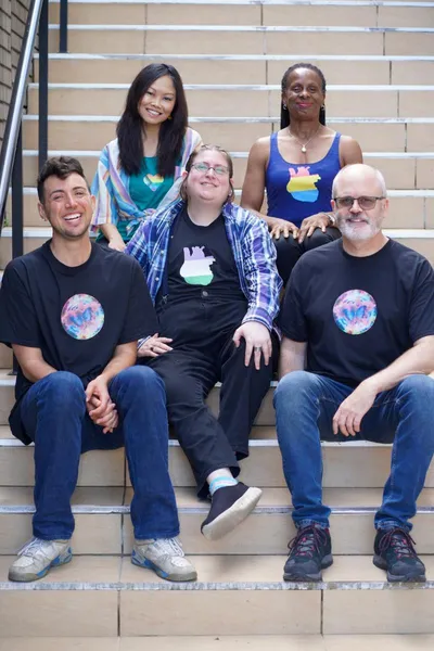 Five people seated in tree rows on concrete stairs. Top row:  An asian woman in a teal tank top glitter printed with a Progress Pride flag in the shape of a heart and a black woman in a purple tank top glitter printed with a pansexual flag in the shape of an anatomical heart. Middle row: A white person in a black t-shirt glitter printed with a genderqueer flag in the shape of an anatomical heart. Bottom row: Two white men both in black t-shirts glitter printed with a circular image of technicolor clouds with a blue butterfly in the center.