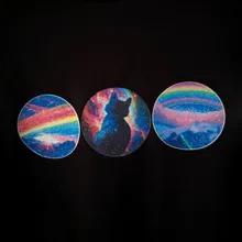 Close up of three circular images arranged in a slightly upward angling line, printed across a black t-shirt. The left and right images show different variations of rainbows over mountain tops, while the middle image show a rear-view of a seated black cat in front of a multicolored nebula