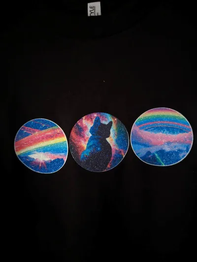 Close up of three circular images arranged in a slightly upward angling line, printed across a black t-shirt. The left and right images show different variations of rainbows over mountain tops, while the middle image show a rear-view of a seated black cat in front of a multicolored nebula