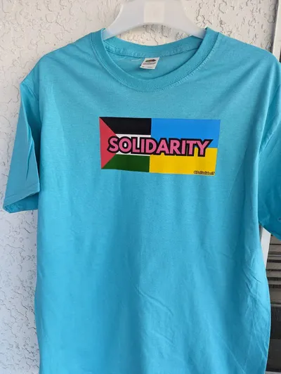 The flags of Palestine and Ukraine next to each other with the word solidarity written across them in pink letters, screen printed on a blue t-shirt.